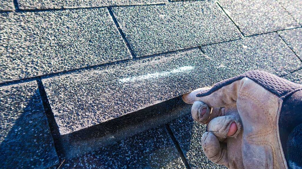 A residential roofer inspects a pitched roof covered with asphalt shingles on a leafy suburban street.