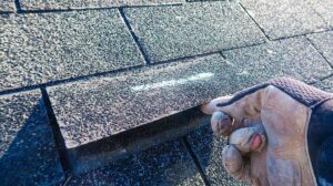 A residential roofer inspects a pitched roof covered with asphalt shingles on a leafy suburban street.