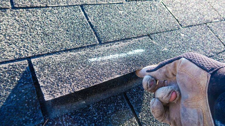 A residential roofer inspects a pitched roof covered with asphalt shingles on a leafy suburban street.